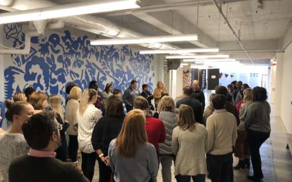 A group of people stand in a semi-circle in a modern office hallway, listening to a speaker. The space features exposed ceilings and blue patterned wall art.