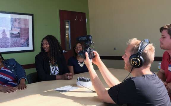 Four people sit around a table, three women on one side smiling and laughing, two men on the other side recording them with a microphone and headphones in a well-lit room with green and yellow walls.