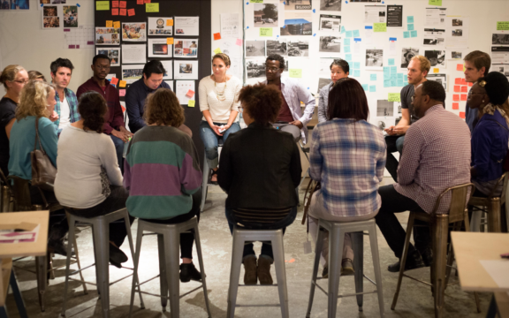 A diverse group of people sit in a circle on stools, engaged in discussion in a bright room with design materials, photos, and sticky notes on the walls behind them.