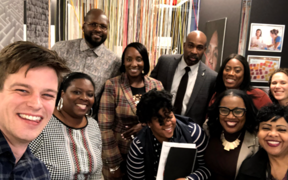 A group of ten people, dressed in business and casual attire, smile together for a selfie in an office setting with colorful string decorations and artwork on the walls.