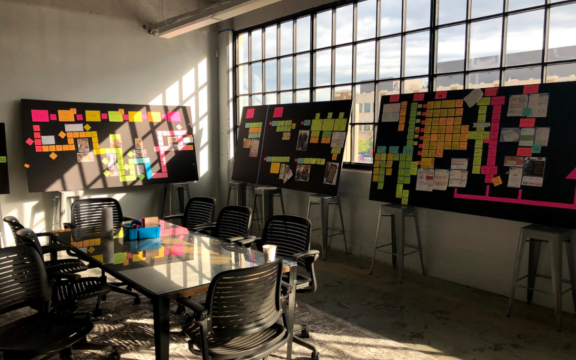 A conference room with a long glass table, black chairs, and three large boards covered with colorful sticky notes, papers, and photos, illuminated by sunlight through tall windows.