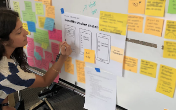 A woman writes on a paper labeled “benefits tracker sketch” attached to a whiteboard covered with colorful sticky notes and diagrams in an office setting.