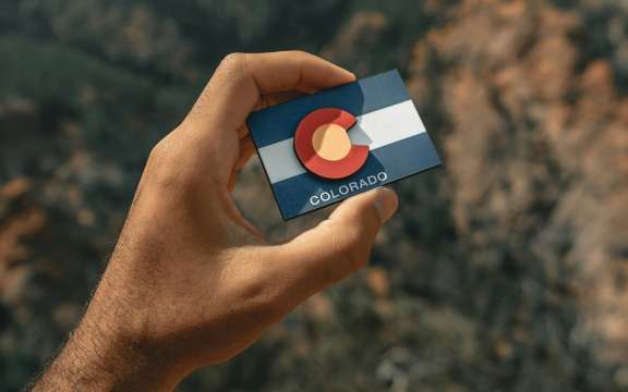 A hand holds a small card featuring the Colorado state flag against a blurred outdoor background of rocky terrain and greenery.