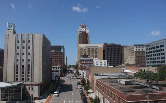 A cityscape view featuring a street lined with buildings, including a tall clock tower in the center under a clear blue sky with a few clouds. Cars are parked along the street, and trees are visible on the sidewalks.