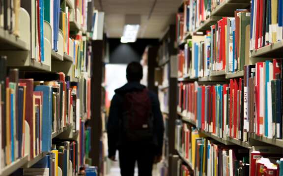 A person with a backpack walks down an aisle between tall bookshelves filled with colorful books in a library, facing away from the camera toward a bright light in the distance.