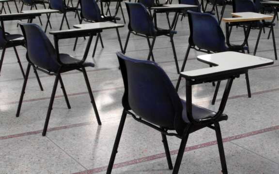 Rows of empty black chairs with small attached writing desks are arranged in a large room, likely set up for an exam or classroom setting. The floor is tiled, and the room appears unoccupied.