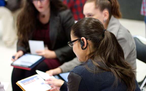 A woman with a hearing aid reads a document while sitting with others in a group setting. The group appears to be engaged in discussion, with tablets and papers in hand.