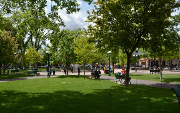 A sunny park scene with people sitting on benches and walking along pathways, surrounded by lush green trees and grass. Some buildings are visible in the background under a partly cloudy sky.