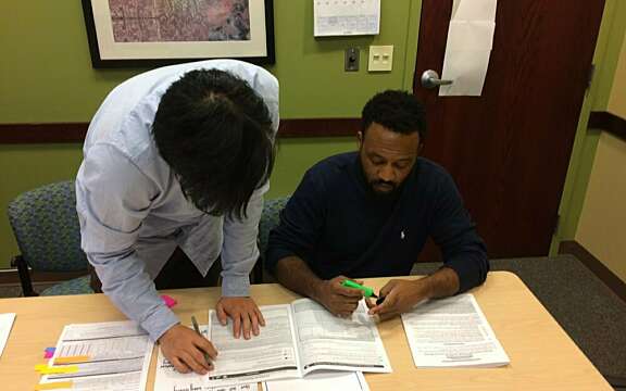 Two people work together at a table covered with papers and highlighters. One person is sitting and highlighting a document, while the other is standing and writing notes. They appear to be collaborating or studying.