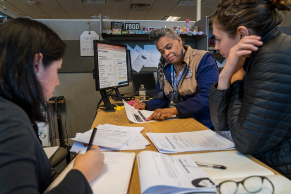Three women sit at a desk in an office, reviewing paperwork together. One woman in a beige vest points to a document while the others take notes. Computer monitors and office supplies are visible in the background.