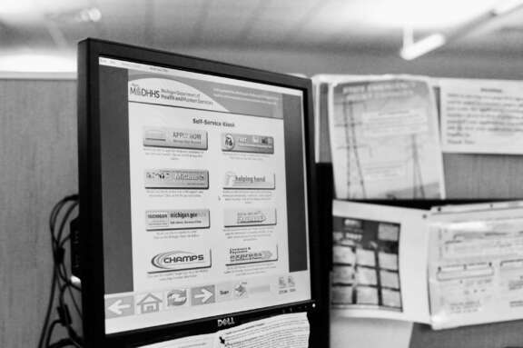 A computer monitor at a desk displays a Michigan Department of Health and Human Services self-service kiosk screen, with various application options and information. Nearby, papers are pinned to a partition wall.