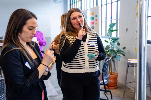 Two women stand indoors near large windows. One writes on a notepad, while the other holds a sticky note and pen thoughtfully to her lips. Bright sticky notes are visible on the wall, and a potted plant is in the background.