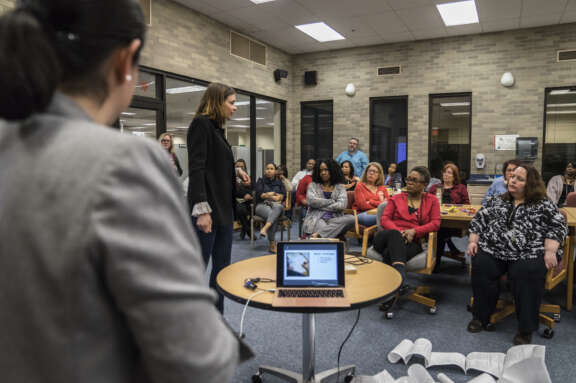 A group of adults sit and listen attentively to a woman speaking in a classroom or meeting room. A laptop displaying a presentation is on a small table in the foreground.