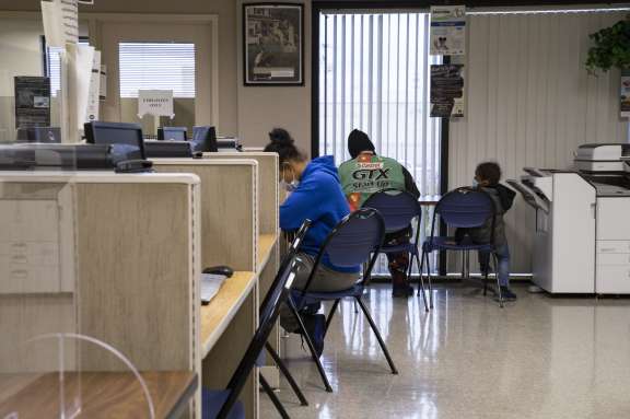 Several people sit spaced apart at desks and chairs in an office or waiting area. Some are writing or working, while a child sits on a chair near a copier or printer. The room has fluorescent lighting and office furniture.