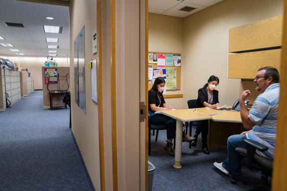 Three people sit around a table in an office, two women wearing masks and a man gesturing while speaking. The room has bulletin boards and visible office spaces in the background.