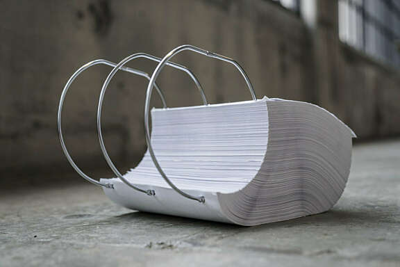 A large stack of paper punched with three holes lies on the ground, held together by oversized metal binder rings. The background is a concrete floor and blurred industrial windows.
