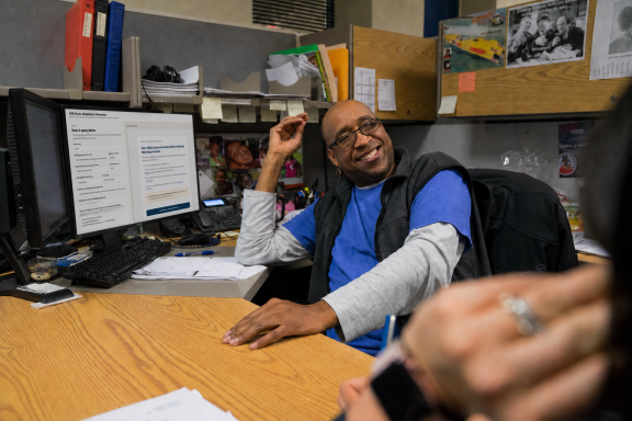 A smiling man in a blue shirt and glasses sits at a cluttered office desk, gesturing with his hand. A computer monitor displays text, and shelves with files and papers are visible in the background.
