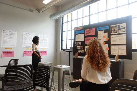 Two people in a brightly lit room view a large presentation board with various papers and images attached. Empty chairs are arranged nearby, and large windows let in natural light.