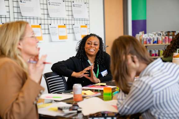 Three women sit around a table covered with papers and colorful notes, engaging in a lively discussion. One woman in the center smiles, while another gestures with her hands. Wall displays and supplies are visible in the background.