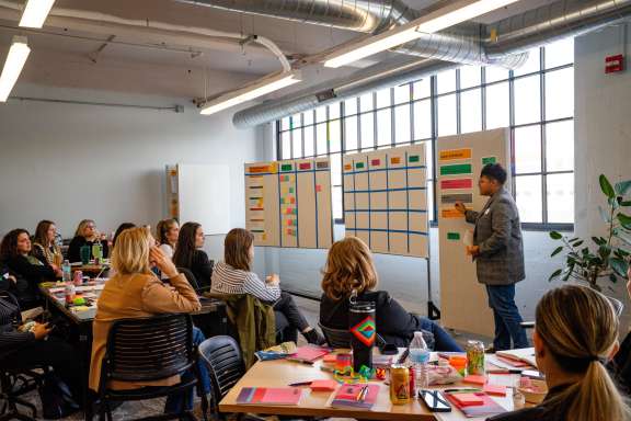 A person stands by whiteboards covered in sticky notes and charts, presenting to a group of seated people in a brightly lit office space with large windows and industrial decor.