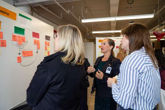 A group of women stands in front of a whiteboard covered with colorful sticky notes and diagrams, discussing ideas in a collaborative office setting.