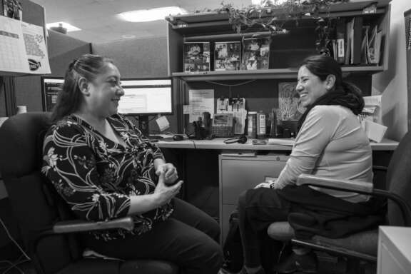 Two women sit facing each other in an office cubicle, smiling and having a friendly conversation. Computer monitors, office supplies, and personal photos are visible on the desk and bulletin board behind them.