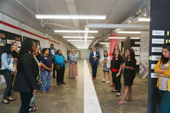A group of people stand in a semi-circle in an industrial-style hallway in Civilla's studio, listening to a speaker. There is a long sheet of paper with diagrams or notes on the floor and posters on the walls.