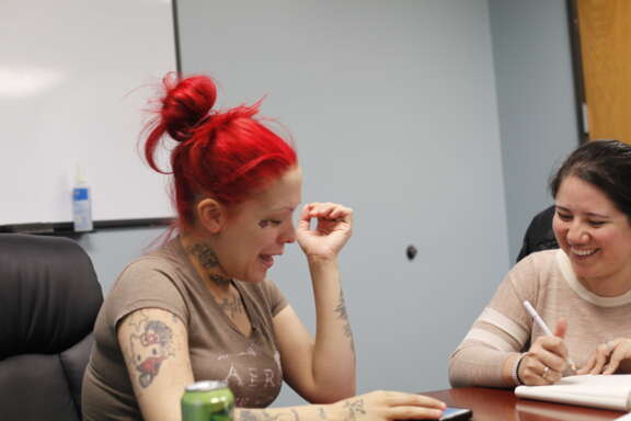 Two women sit at a table, smiling and talking. One has bright red hair in a messy bun and visible tattoos, while the other writes in a notebook. A whiteboard and soda can are in the background.