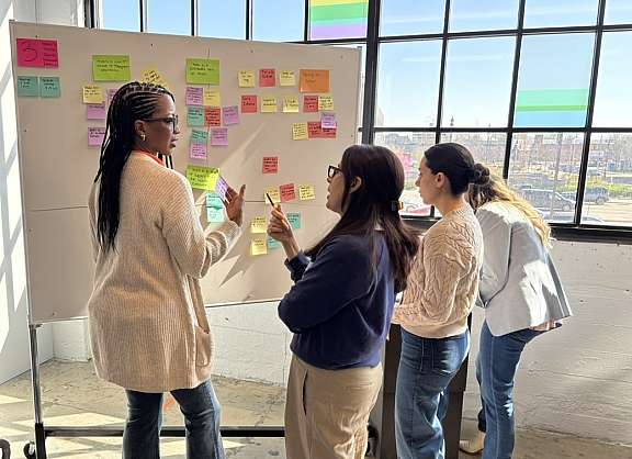 Three women stand by a large window, discussing colorful sticky notes on a whiteboard. Sunlight streams in, creating a bright atmosphere in the modern office space.