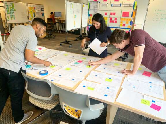 Three people stand around a table covered with papers and colorful sticky notes, collaborating and reviewing documents in a modern office with whiteboards and charts in the background.