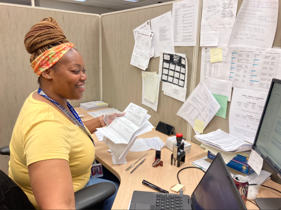 A woman in a yellow shirt sits at a desk in a cubicle, smiling while sorting papers. The desk and walls are covered with documents, notes, and a calendar. She is working on a laptop, with office supplies and a soda can nearby.