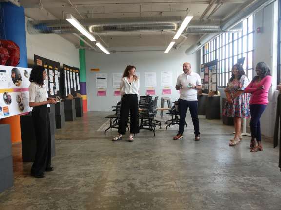 A group of six people stands in a brightly lit room in Civilla's studio with large windows, listening to a woman presenting next to display boards with images and text. Empty chairs and posters are visible in the background.
