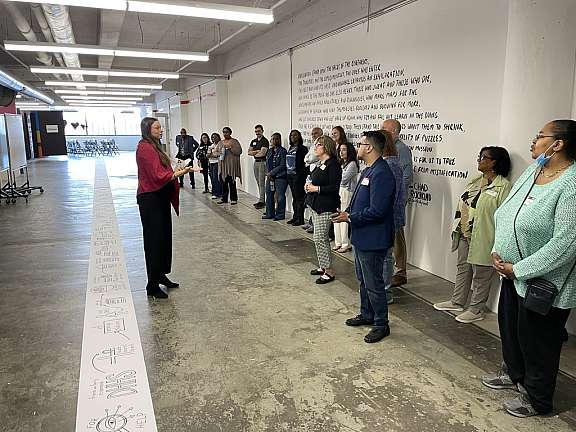 A person speaks to a diverse group of people standing in a semi-circle inside a spacious, industrial-style room with wall art and handwritten text on the wall behind them.