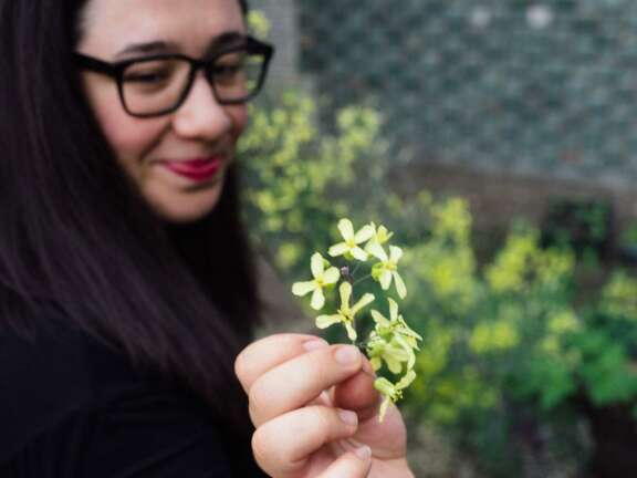 A woman with long dark hair and glasses smiles softly while holding a small bunch of yellow-green flowers in her hand, with a blurred garden background.