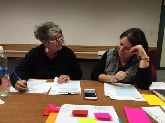 Two women sit at a table with papers, pens, and colorful sticky notes. One woman wears glasses and large earrings, and they appear to be discussing or working on documents together. A water bottle and a phone are on the table.