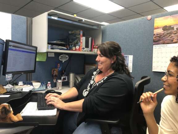 Two women sit at a desk in an office cubicle, one typing on a keyboard and smiling at a computer monitor, while the other holds a pen and looks on. Office supplies and a November calendar are visible in the background.