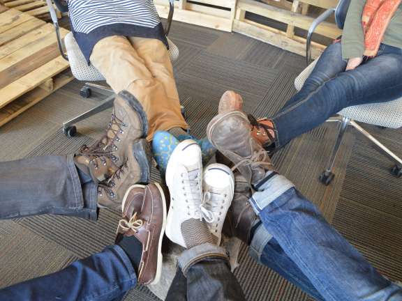 Five people sitting in chairs have stretched their feet toward the center, showing a variety of shoes and pants. The scene appears casual and relaxed, with wooden pallets visible in the background.