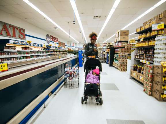 A woman pushes a stroller with a bundled-up child through a grocery store aisle, surrounded by shelves stocked with canned goods and other products. Bright store lighting and signs are visible overhead.