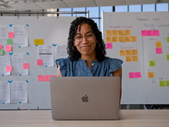 A woman with curly hair and glasses smiles while working on a laptop at a desk, with whiteboards covered in colorful sticky notes and papers in the background.