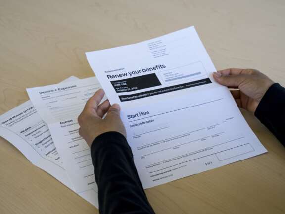 A person holding paperwork titled Renew your benefits at a table, with additional documents labeled “Income + Expenses” beneath their hands. The forms appear to be related to financial or social services.