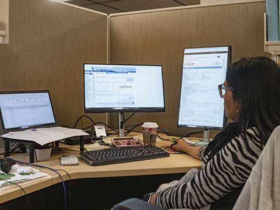 A person sits at a desk in an office cubicle, working on two large computer monitors with spreadsheets and emails open. Papers, a keyboard, headphones, a coffee cup, and office supplies are scattered on the desk.