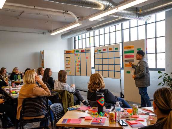 A person stands by whiteboards covered in sticky notes and charts, presenting to a group of seated people in a brightly lit office space with large windows and industrial decor.