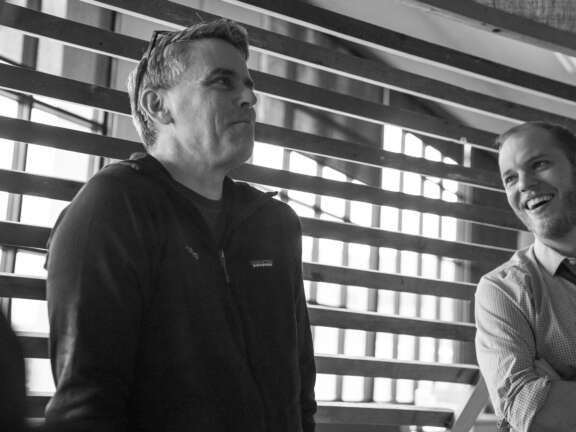 Two men stand indoors by a window with wooden slats. One man in a jacket looks thoughtful, while the other, wearing a shirt and tie, smiles and laughs. The image is in black and white.