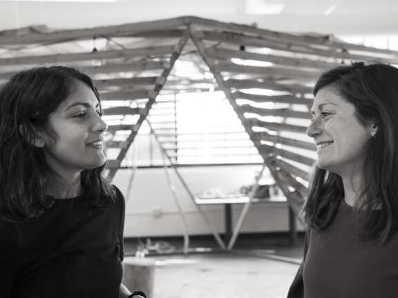 Two women stand indoors, facing each other and smiling, in front of a wooden, geometric structure. The image is in black and white, and the setting appears to be a creative or collaborative workspace.
