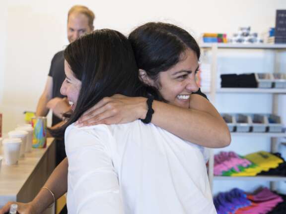 Two women smiling and hugging in a bright room with shelves of colorful t-shirts and books in the background; a man stands nearby, slightly out of focus.