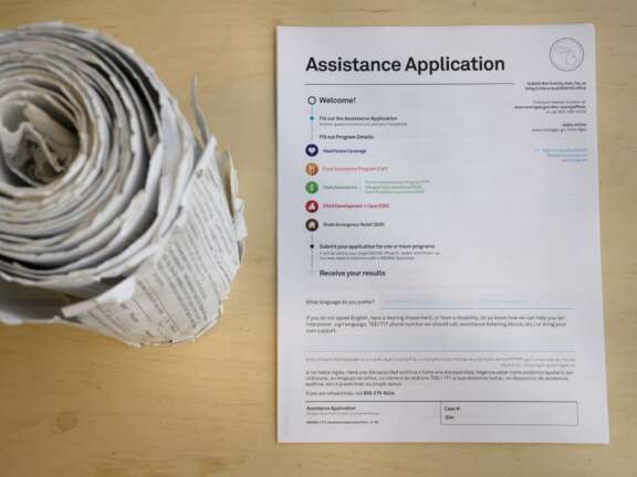 A rolled stack of papers sits on a wooden table next to an assistance application form with colorful icons and text instructions for submitting and receiving results.