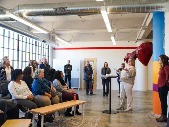 A group of people sit and stand in a brightly lit room, attentively listening to a woman speaking at a podium. Posters are displayed on the wall and a large red sculpture is in the background.