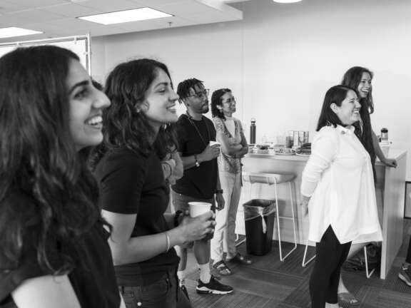 A group of people standing in a room, smiling and watching something off camera. Some hold cups, and there are tables and chairs in the background. The mood appears relaxed and cheerful. The image is in black and white.