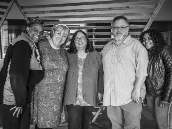 Five adults, three women and two men, stand closely together smiling for the camera in a casual indoor setting, with wooden beams in the background. The image is in black and white.