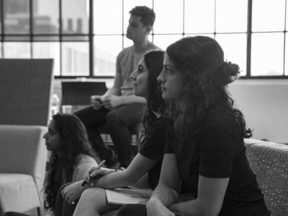 A group of four people, three women and one man, sit attentively in a bright room with large windows, listening and taking notes during a meeting or lecture. The image is in black and white.